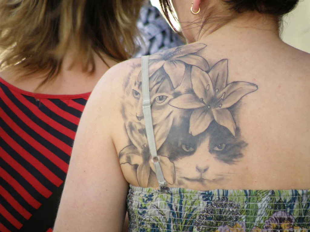 50 Animal Tattoo Ideas: The Complete Guide by Animal, Style and Placement 25 Black and grey cat tattoo partly hidden behind large lily flowers on a shoulder blade