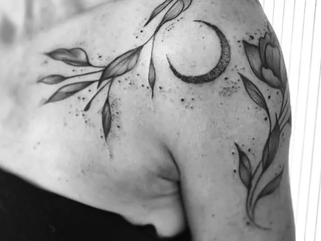 25 Fine Line Tattoo Ideas: Delicate Designs, Honestly Rated 4 Black and white photo of a fine line leafy stem tattoo with a crescent moon on a shoulder