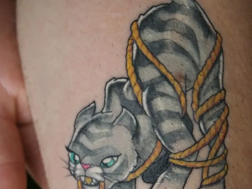 22 Pet Tattoo Ideas: Portraits, Paw Prints and Memorials Done Right 13 Colour tattoo of a tabby cat tangled in yellow yarn on an arm