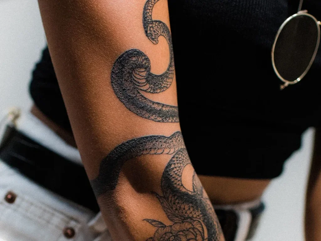 20 Snake Tattoo Ideas: Meanings, Styles and Placements That Work 6 Blackwork snake tattoo winding through peonies down a full arm